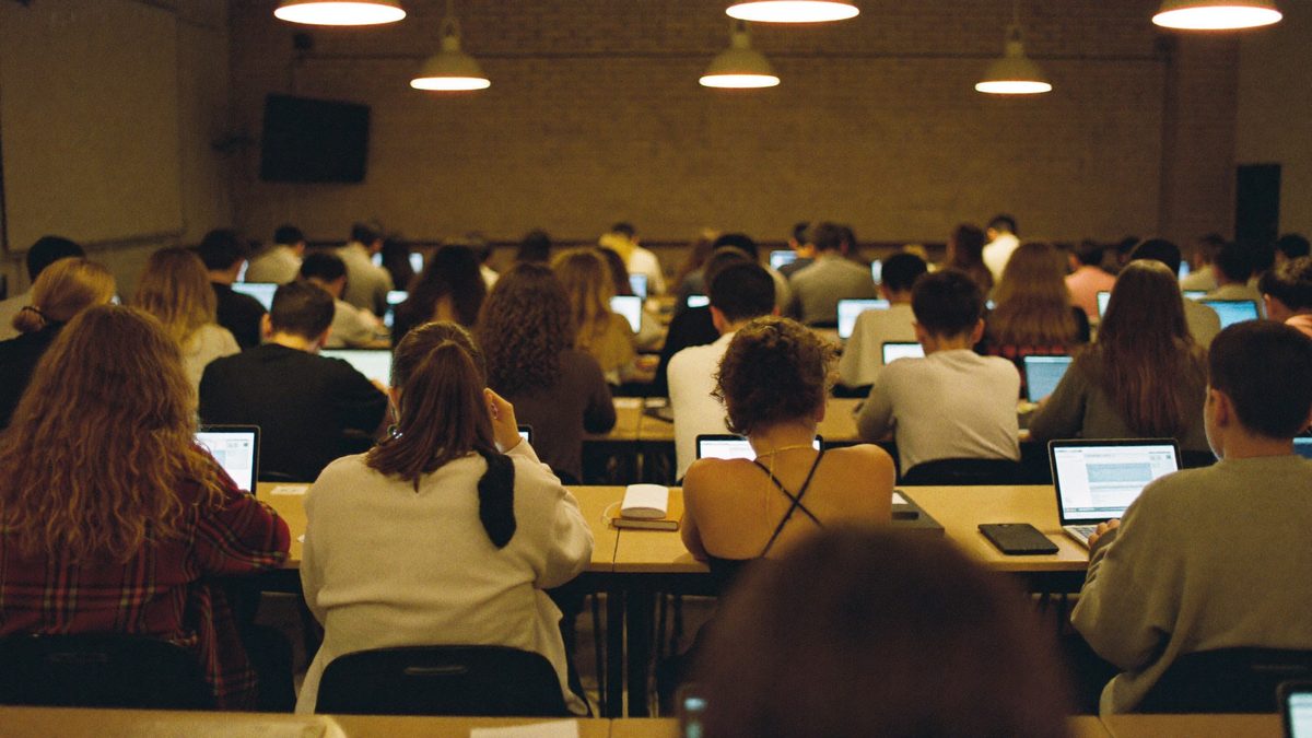 Students working on laptops in a warm classroom, seen from the back of the room
